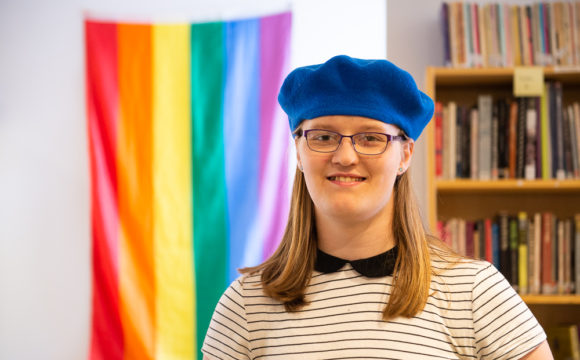 A photo of Penelope in a library standing in front of a rainbow flag wearing a blue beret.